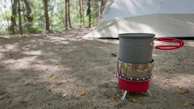 A measured metal camping mug sits on a red portable stove at a quiet forest campsite. Sunlight filters through surrounding trees onto the compact outdoor kitchen gear near a tent.