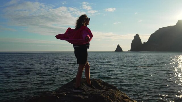 Woman sea sunset standing on a coastal rock with jacket blowing in the wind while enjoying the peaceful evening landscape