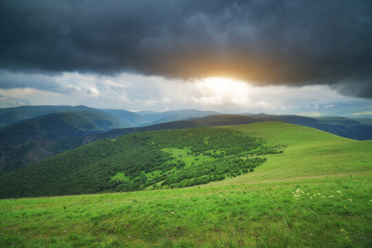 Bright sunset breaks through dark storm clouds over lush green hills and alpine meadows. Majestic mountain landscape