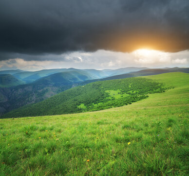 Bright sunset breaks through dark storm clouds over lush green hills and alpine meadows. Majestic mountain landscape