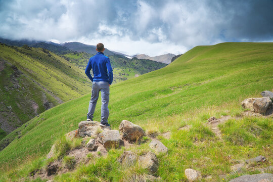 Man in a blue stands on rocks looking at a vast mountain valley with green slopes under cloudy sky. Back view tracking concept
