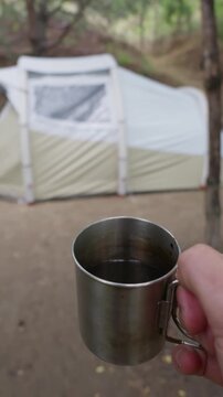 A close-up view of a person holding a stainless steel metal mug filled with hot dark coffee at a quiet campsite. In the out-of-focus background, a canvas camping tent stands among trees.