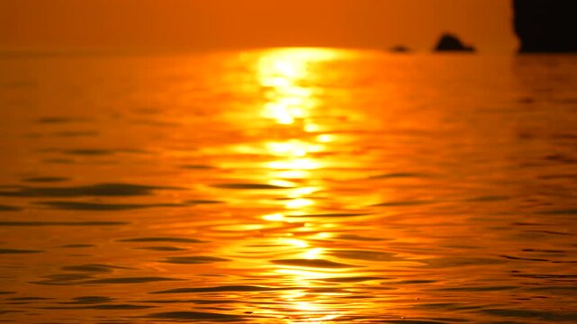 Golden light glimmers across gentle sea ripples at sunset near a rocky coastline, providing a serene atmosphere. Eye level static shot.