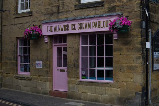 Quaint pink wooden storefront of The Alnwick Ice Cream Parlour set in a historic rustic stone building with hanging flower baskets in England.