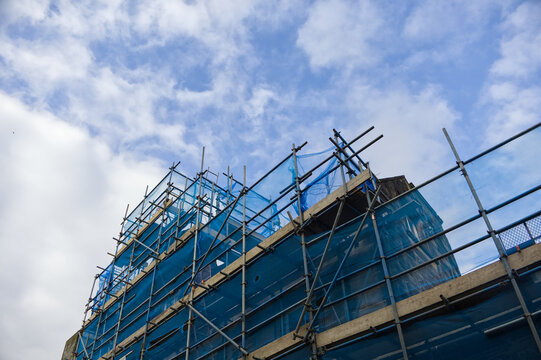 Construction site with metal scaffolding and blue safety netting on a building exterior against a partly cloudy sky, urban development concept.