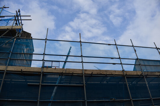 Construction site with metal scaffolding and blue safety netting on a building exterior against a partly cloudy sky, urban development concept.