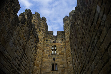 Majestic medieval stone castle fortress featuring ancient defense walls, battlements, and tall towers surrounded by green grass under a cloudy sky. Alnwick castle England, towers a very old castle © TomKorcak