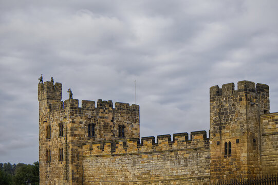 Majestic medieval stone castle fortress featuring ancient defense walls, battlements, and tall towers surrounded by green grass under a cloudy sky. Alnwick castle England, towers a very old castle