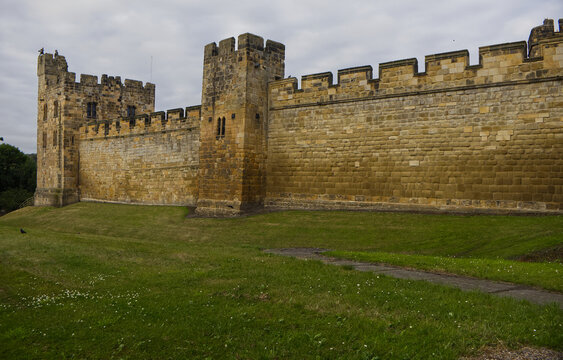 Majestic medieval stone castle fortress featuring ancient defense walls, battlements, and tall towers surrounded by green grass under a cloudy sky. Alnwick castle England, towers a very old castle