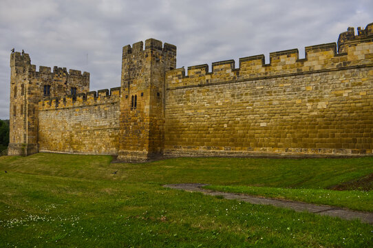 Majestic medieval stone castle fortress featuring ancient defense walls, battlements, and tall towers surrounded by green grass under a cloudy sky. Alnwick castle England, towers a very old castle