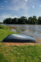 Upside-down small boat rests on a grassy lakeshore beside calm water, reeds, trees, and a paved launch area under a bright blue summer sky with copy space © ugis