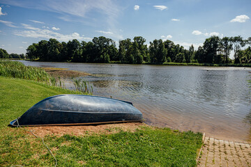 Upside-down small boat rests on a grassy lakeshore beside calm water, reeds, trees, and a paved launch area under a bright blue summer sky with copy space © ugis