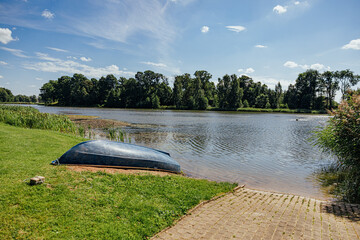 Upside-down small boat rests on a grassy lakeshore beside calm water, reeds, trees, and a paved launch area under a bright blue summer sky with copy space © ugis