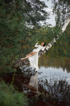 person dressed in white reclines on a tree branch over water. scene shows green foliage and a calm environment on a clear day. Reflections are visible in the water