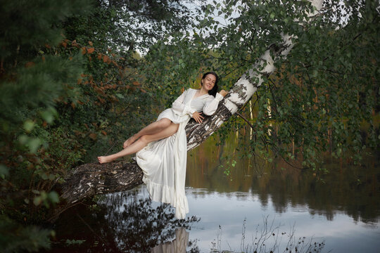 woman in a white dress is lying on a tree branch that hangs over a body of water. setting is a green forest with trees and calm water. It is daytime and sunny