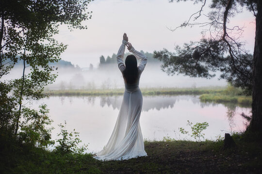 woman in a white dress stands near a lake at dawn. Her hands are raised, and fog surrounds the area. Trees frame the scene with soft light in the background