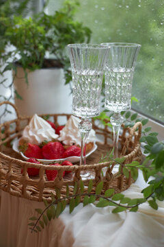 Two crystal glasses sit on a woven tray with strawberries and meringue beside them. The window shows green plants outside, creating a cozy atmosphere for enjoying a treat