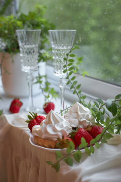 A plate of dessert with strawberries and cream sits on a table near a window. Two tall glasses stand beside the plate. Green leaves decorate the table setting