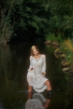 A model stands in a river wearing a crochet dress. The sun shines through the trees, creating a bright scene. Water reflects the models figure as she poses