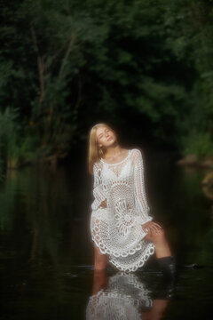 A model stands in shallow water wearing a white dress made of crochet. The scene takes place in a forested location with greenery all around under bright sunlight