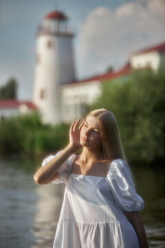 Woman stands by the river, touching her face as she looks away from the camera. A lighthouse is visible behind her, and trees frame the scene in the background