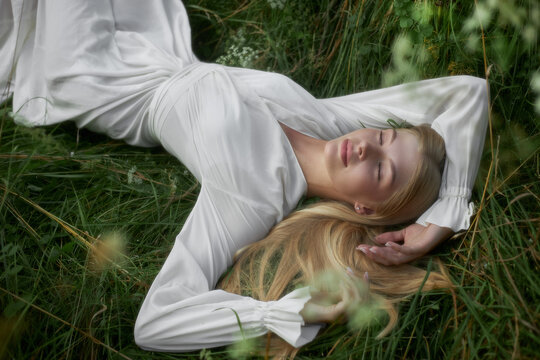 A woman rests on green grass in a field. She wears a white dress and has long hair spread around her. Sunlight shines down while she enjoys the moment outdoors