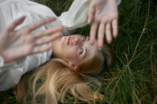A girl is lying on the grass with her hands raised up. She has long hair and is wearing a white shirt. Sunlight shines on her face. The scene is calm and natural