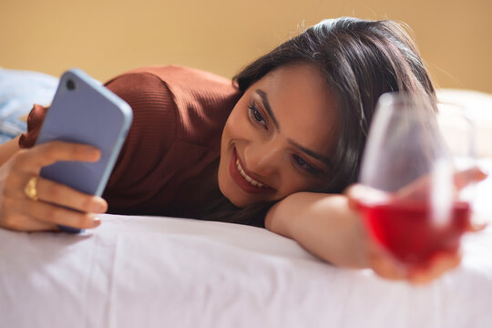 Young woman using Smartphone with holding a glass of wine while lying down on bed