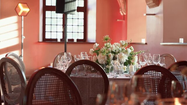 Beautifully decorated table set with glasses and white floral centerpiece, sunlight streaming through vintage window, warm ambient lighting, elegant romantic wedding venue, nobody visible.