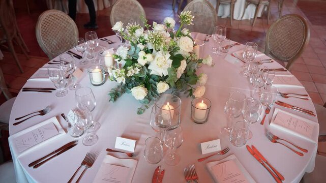 Beautifully decorated round table set for a wedding reception, featuring white floral centerpiece, glowing candles, and fine tableware in soft, ambient lighting. Nobody present.