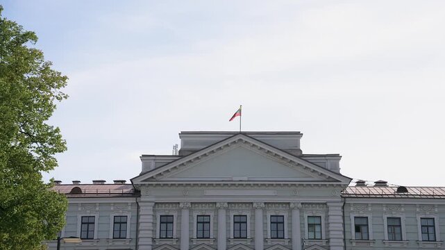 Stately neoclassical Vilnius Regional Court facade in Lithuania, featuring the national flag atop the roof. Bright daylight, clear sky, and lush tree frame the elegant architecture.