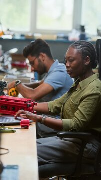 Vertical video of African American young adult woman programming robotic vehicle on laptop in workshop. Clip is ideal for STEM education and robotics engineering promotional campaigns