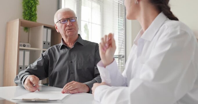 Senior man consultant reviews agreement documents with advisor. A client and woman advisor discuss terms at office desk, reviewing paperwork for a major decision. Clear support for later life.