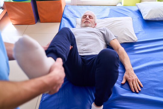 Senior Man Receiving Leg Physiotherapy From Therapist On Blue Mat Indoors For Injury Rehabilitation And Improved Mobility After Surgery