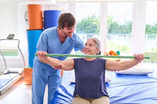 Senior Woman Doing Rehabilitation Exercises With Physiotherapist Using Bar In Rehab Center For Recovery And Improved Mobility Indoors