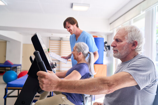 Senior Man Exercising On Stationary Bike With Physiotherapist Assisting In Rehabilitation Center For Recovery And Improved Physical Fitness