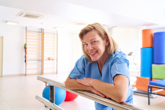 Smiling Physiotherapist Leaning On Parallel Bars In Rehabilitation Center For Physical Therapy And Recovery After Injury Indoors