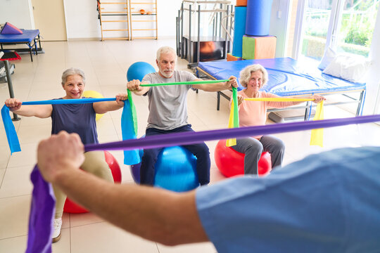 Senior Adults Exercising With Resistance Bands On Exercise Balls In Rehabilitation Center With Physiotherapist Supervision For Recovery
