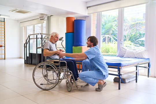 Senior Man In Wheelchair Assisted By Physiotherapist During Rehabilitation Session Indoors At Rehab Center For Recovery And Improved Mobility