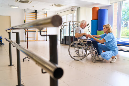 Senior Man In Wheelchair Assisted By Physiotherapist During Rehabilitation Session Indoors At Rehab Center For Recovery And Improved Mobility