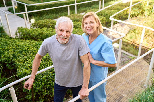Senior Man With Cane Walking Up Steps Assisted By Physiotherapist Outdoors In Sunlight During Rehabilitation Session For Recovery