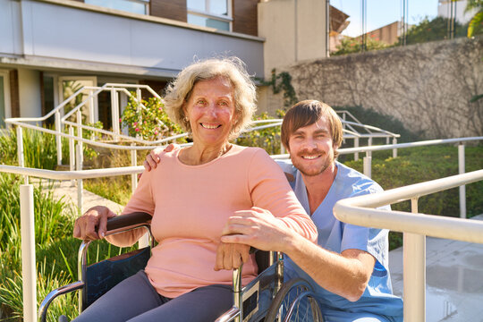 Senior Woman in Wheelchair with Physiotherapist Outdoors at Rehab Center Smiling and Receiving Care and Support During Rehabilitation Process