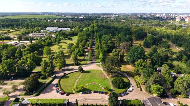 Aerial view over Herrenkrug street and Elbauenpark in Magdeburg, with landscaped lawns, tree-lined avenues and city skyline. Summer panorama shows green parkland, Elbe corridor and urban surroundings