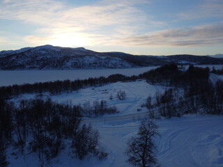 Aerial drone view of a serene snow-covered mountain landscape and frozen lake during a peaceful winter sunset in the remote Arctic wilderness. © nikjuzaili