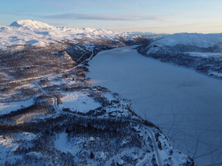 Stunning aerial perspective of a vast frozen lake surrounded by snow-covered mountains and winding roads in a Nordic winter landscape at sunrise © nikjuzaili