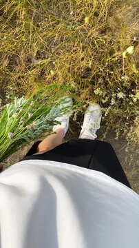 A woman s legs in the grass and a bouquet of wildflowers in her hand, viewed from above. Unity with nature, rest, relaxation, enjoying the summer atmosphere, outdoor recreation, hiking