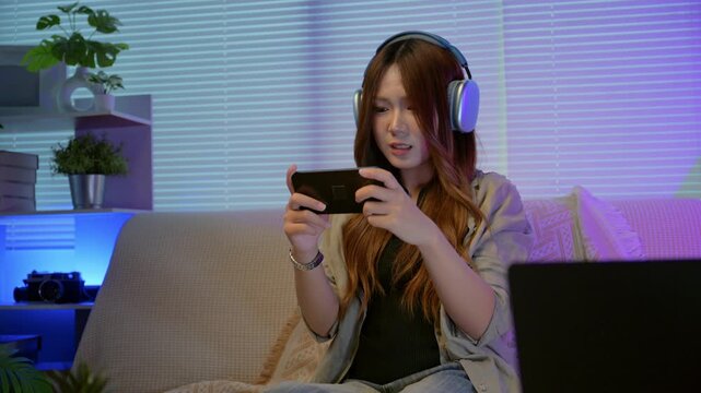 Young Asian gamer presses on her smartphone screen to stream exciting and fun gameplay at home amidst a warm, neon-lit living room. Concept of mobile gaming lifestyle, modern digital entertainment.