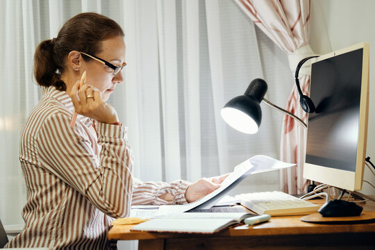Mid-adult woman in glasses turning pages of documents at home desk with lamp and computer monitor while working on project in evening.