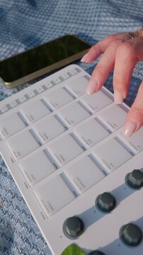 A close-up view of a woman hands playing beats on a white MIDI drum pad controller. The female musician creates electronic music outdoors while relaxing on a blanket in the bright sunlight.