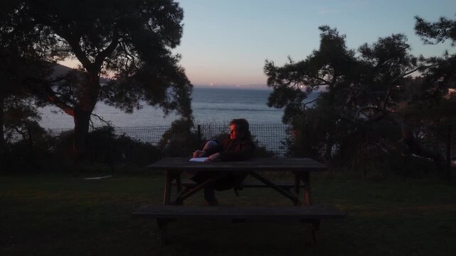 Thoughtful young man sitting at wooden bench table holding notebook looking at sunset ocean view cinematic wide shot
